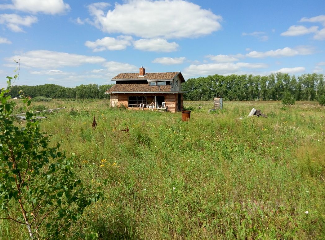Дом в загородном стиле с деревянной отделкой и крышей из черепицы. Фасад имеет большие окна, обеспечивающие естественное освещение. Перед домом расположена веранда с несколькими стульями, что создает уютную атмосферу. Стены выполнены из бревенчатого материала, что подчеркивает природный стиль. Крыша с небольшим наклоном и мансардным окном добавляет характер. Цветовая гамма включает теплые древесные оттенки, создавая гармонию с окружающей природой. Пространство вокруг дома не застроено, что придает ощущение уединения и спокойствия.