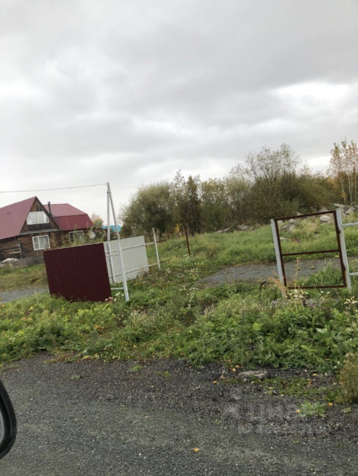 На фотографии представлено загородное строение в традиционном русском стиле с деревянными стенами и крышей из металлочерепицы красного цвета. Видно, что дом имеет два этажа и большие окна, что позволяет естественному свету проникать внутрь. Вокруг дома расположены участки с заросшей травой и кустарниками, а также забор из белых и красных панелей. Площадка перед домом выглядит неухоженной, с каменистым покрытием и несколькими металлическими конструкциями. Общая атмосфера создает ощущение уединенности и близости к природе.