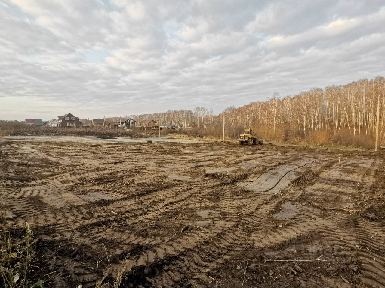 На фотографии представлено пустое земельное пространство с видимыми следами от строительной техники. Поверхность земли рыхлая, с видимыми следами от колес. На заднем плане видны несколько жилых домов, выполненных в традиционном стиле, с деревянными фасадами и крышами, покрытыми черепицей. Пространство выглядит подготовленным для дальнейших строительных работ. Небо затянуто облаками, что создает спокойную атмосферу. Общая композиция изображения передает ощущение начального этапа развития территории.