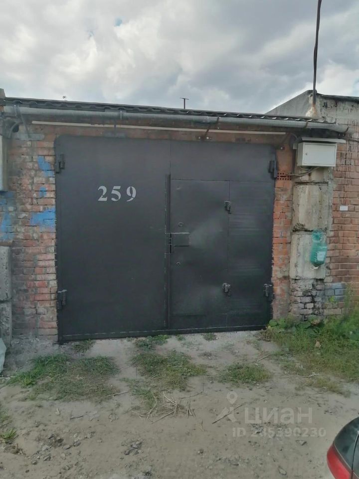Видна металлическая дверь с номером 259, выполненная в индустриальном стиле. Стены гаража сложены из кирпича, некоторые участки имеют неравномерную окраску и следы времени. Дверь имеет простую конструкцию с несколькими замками и петлями. Вокруг двери заметны следы от использования, а также элементы, указывающие на функциональность помещения. Пол перед гаражом покрыт землёй и травой, что подчеркивает его утилитарное назначение. Общая цветовая гамма включает серые и коричневые оттенки, создающие ощущение прочности и надежности.
