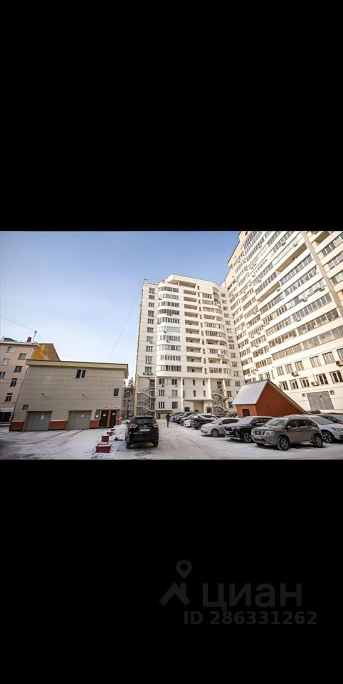 Современное многоквартирное здание с характерными элементами архитектурного стиля, такими как большие панорамные окна и балконы. Видна структура с несколькими этажами, где дом имеет прямые линии и острые углы. Внутренний двор оформлен с парковочными местами для автомобилей. Основные материалы фасада — бетон и стекло, с нейтральными цветами, такими как белый и серый. Дворовая территория аккуратно оформлена, с минималистичными элементами, создающими современный городской стиль.