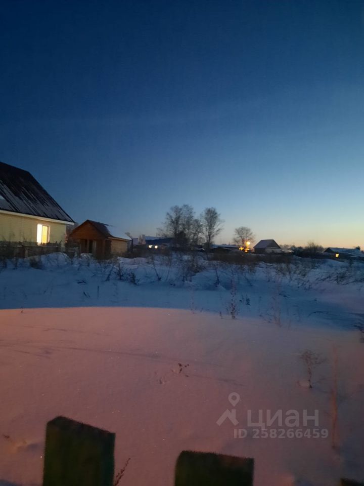 На фотографии виден зимний пейзаж с заснеженной территорией. В центре кадра расположены следы на снегу, указывающие на активность. Слева виден дом с крышей, покрытой темным материалом, и окнами, светящимися в вечернем свете. На заднем плане можно увидеть несколько строений с крышей, также покрытой снегом. Цветовая гамма включает холодные оттенки синего и белого, создавая атмосферу спокойствия. В целом, пространство выглядит уединенным и тихим, характерным для зимней природы.