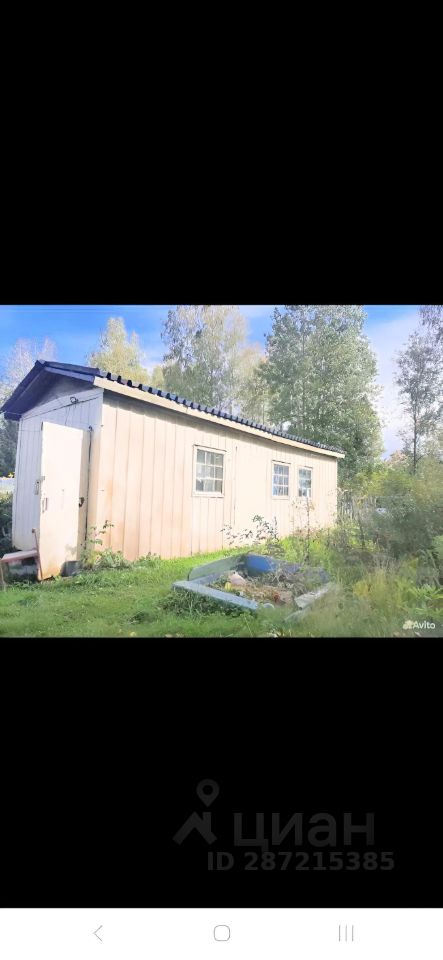 Строение выполнено в простом, функциональном стиле с деревянными стенами и двускатной крышей. Видны окна с рамами, а также входная дверь, расположенная сбоку. Конструкция имеет прямоугольную форму и небольшие размеры, что делает её подходящей для использования в качестве дачи или хозяйственного помещения. Цвет стен светлый, возможно, с использованием краски или природного дерева. Отсутствие сложных архитектурных деталей подчеркивает практичность и функциональность постройки.