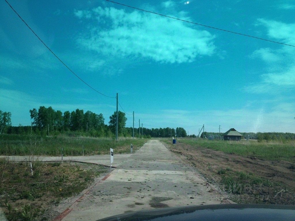 На фотографии виден участок земли с неасфальтированной дорогой, окружённый зелёными деревьями и травой. Дорога имеет бетонные плиты, которые частично покрыты грязью. Справа расположено небольшое здание с крышей, вероятно, дача или хозяйственное строение. Вдали видны линии электропередач. Общая атмосфера участка спокойная и уединённая, что создаёт ощущение загородной жизни.