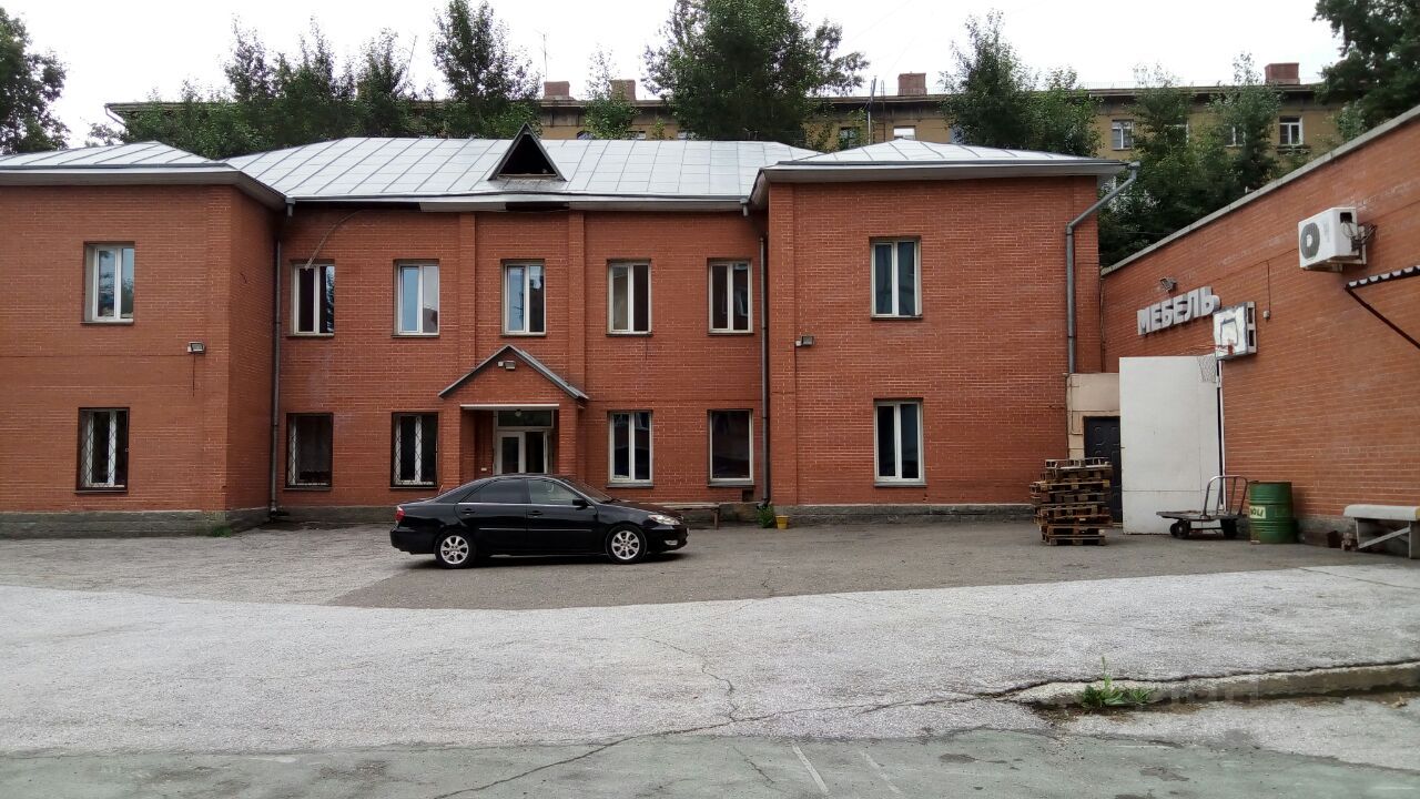 Здание выполнено в стиле промышленной архитектуры с красным кирпичным фасадом и металлической крышей. На переднем плане виден черный автомобиль, стоящий на асфальтированной площадке. Окна прямоугольной формы с простыми рамами, что придает зданию строгий вид. Вход оформлен крытой лестницей, что добавляет функциональности. На одной стороне здания расположена вывеска с надписью 'Мебель', указывающая на назначение помещения. Стены отделаны кирпичом, а крыша имеет современное покрытие, что подчеркивает практичность и долговечность конструкции.