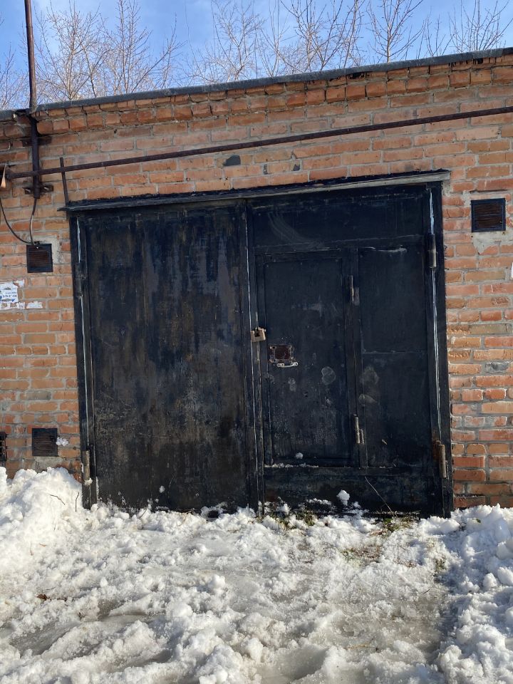 Кирпичная стена с грубой текстурой, выполненная из красного кирпича, создает индустриальный стиль. На переднем плане расположены массивные металлические двери черного цвета, которые имеют заметные следы коррозии и износа, что придает им характерный вид. Двери оснащены замком и дополнительной дверцей, что указывает на функциональность. Внизу виден снег, который частично покрывает землю, создавая контраст с теплым цветом кирпича. Отсутствие окон в этой части здания подчеркивает закрытость и защищенность пространства.
