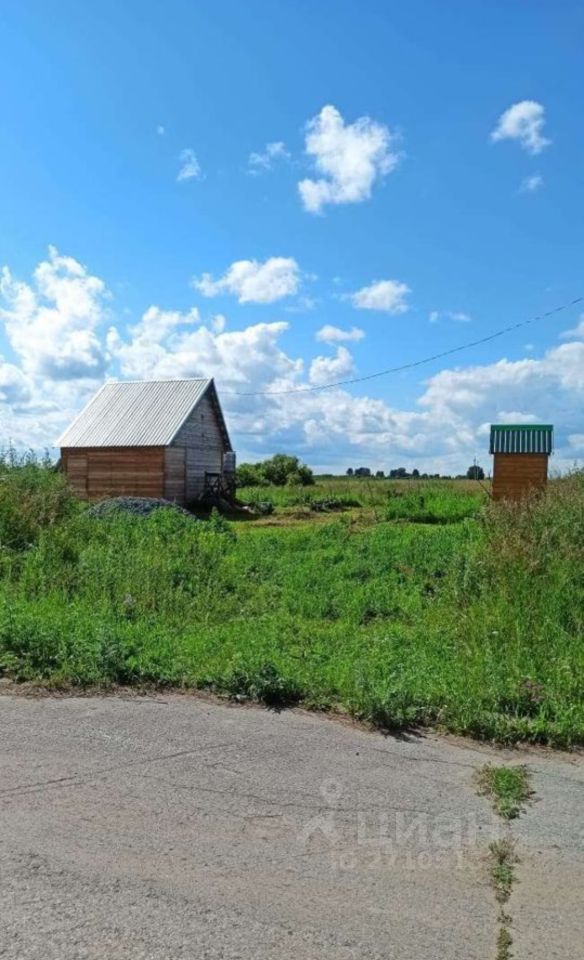 Деревянный дом в стиле загородного жилья с наклонной крышей и металлической черепицей. Стены выполнены из бруса, что придаёт естественный вид. Видна простая, но функциональная планировка с небольшим крыльцом. Рядом расположена постройка с крышей в зелёных тонах, возможно, для хранения. Цвета преобладают натуральные – коричневый, зелёный и серый, что гармонирует с окружающей природой. Пространство вокруг дома не застроено, создавая атмосферу уединения и спокойствия. Общая отделка выглядит простой, но аккуратной, что подчеркивает загородный стиль.