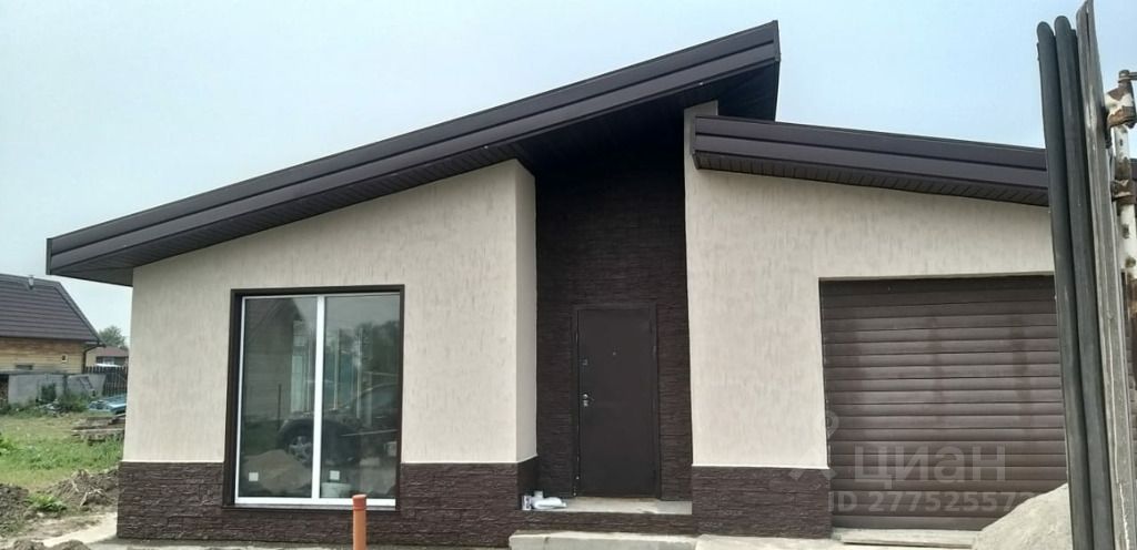 Современный одноэтажный дом с плоской крышей и угловыми элементами. Фасад выполнен в сочетании светлой штукатурки и темного камня, создавая контраст. Большое панорамное окно обеспечивает естественное освещение и визуальную связь с внешним пространством. Входная дверь расположена по центру, рядом с гаражными воротами, что подчеркивает функциональность и удобство. Крыша имеет наклон, что добавляет динамики в архитектурный стиль. Используемые материалы выглядят современно и качественно, а цветовая палитра сочетает нейтральные тона, что делает дом привлекательным и стильным.