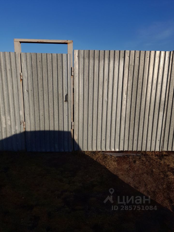 На фотографии виден металлический забор с вертикальными полосами, выполненный из оцинкованного железа. Забор имеет двустворчатые ворота, которые частично открыты, с простым механизмом запирания. Цвет забора серый, с характерными следами коррозии на верхней части ворот. У основания забора видна земля с небольшими участками травы, что указывает на наличие ухоженной территории. Отсутствие дополнительных элементов декора придает забору функциональный и практичный вид, подходящий для загородного участка.