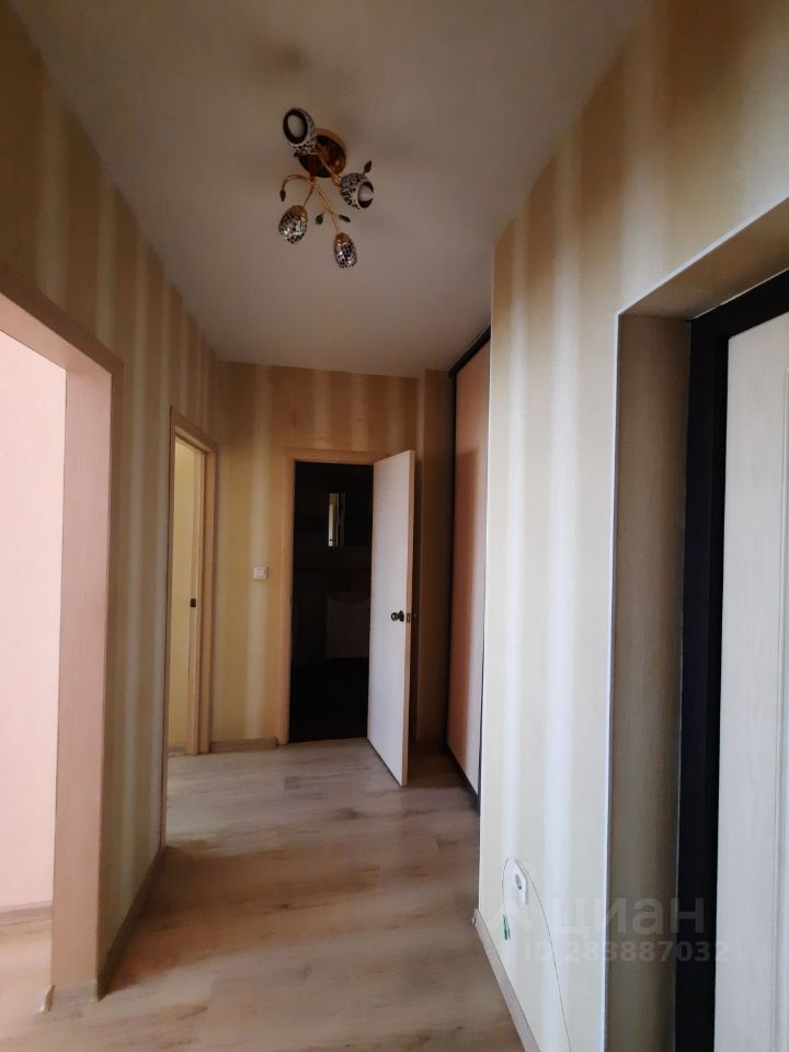 Коридор с современным дизайном, оформленный в светлых тонах. Стены покрыты обоями с вертикальными полосами, создающими эффект высоты. Потолок украшен стильной люстрой с несколькими плафонами. На полу уложен светлый ламинат, который гармонирует с общим интерьером. В коридоре видны двери, ведущие в соседние комнаты, выполненные в светлом дереве. Пространство выглядит уютным и светлым, что создает приятное первое впечатление о доме.