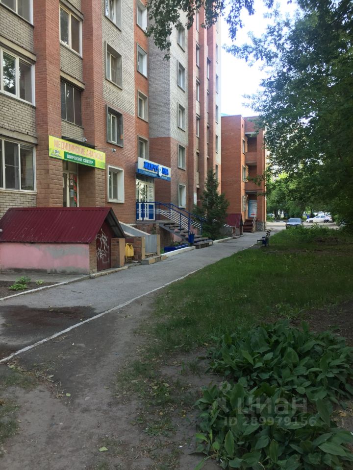 На фотографии представлено здание в современном архитектурном стиле с элементами кирпичной кладки. Стены выполнены в сочетании красного и серого цветов, что придаёт фасаду динамичность. На переднем плане виден пешеходный путь, выложенный асфальтом, который ведёт к входу в здание. Вход оформлен синими металлическими перилами. Установлены окна стандартного размера с простыми рамами. Рядом расположены небольшие хозяйственные постройки с красной крышей, а также зелёные насаждения, создающие уютную атмосферу. Общий вид пространства выглядит аккуратно и организованно.