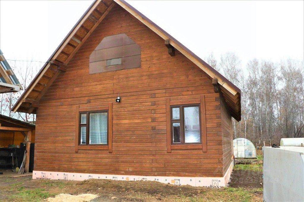 Дом выполнен в традиционном деревянном архитектурном стиле с элементами кантри. Видно, что фасад обшит деревянными панелями, придающими теплый и естественный вид. Структурные особенности включают наклонную крышу с выступающими карнизами и небольшое окно с рамами из дерева. Внешний вид окна украшен деревянными наличниками. Цветовая гамма доминирует в теплых коричневых тонах, что создает уютную атмосферу. Отделка выполнена с использованием натурального дерева, что подчеркивает экологичность и гармонию с природой.