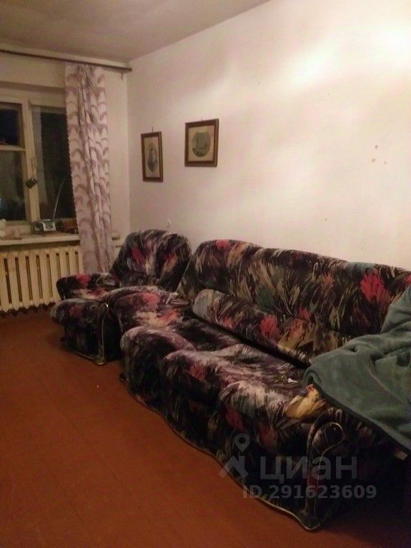 В интерьере видны два дивана с ярким цветным текстилем, создающим уютную атмосферу. На стене расположены две картины в простых рамках, добавляющие художественный элемент. Окно, обрамленное шторами с абстрактным узором, обеспечивает естественное освещение. Пол выложен деревянными панелями, что придает помещению тепло. Видно радиатор отопления под окном, что свидетельствует о функциональности пространства. Общая цветовая палитра включает теплые оттенки, создающие комфортную обстановку для отдыха.