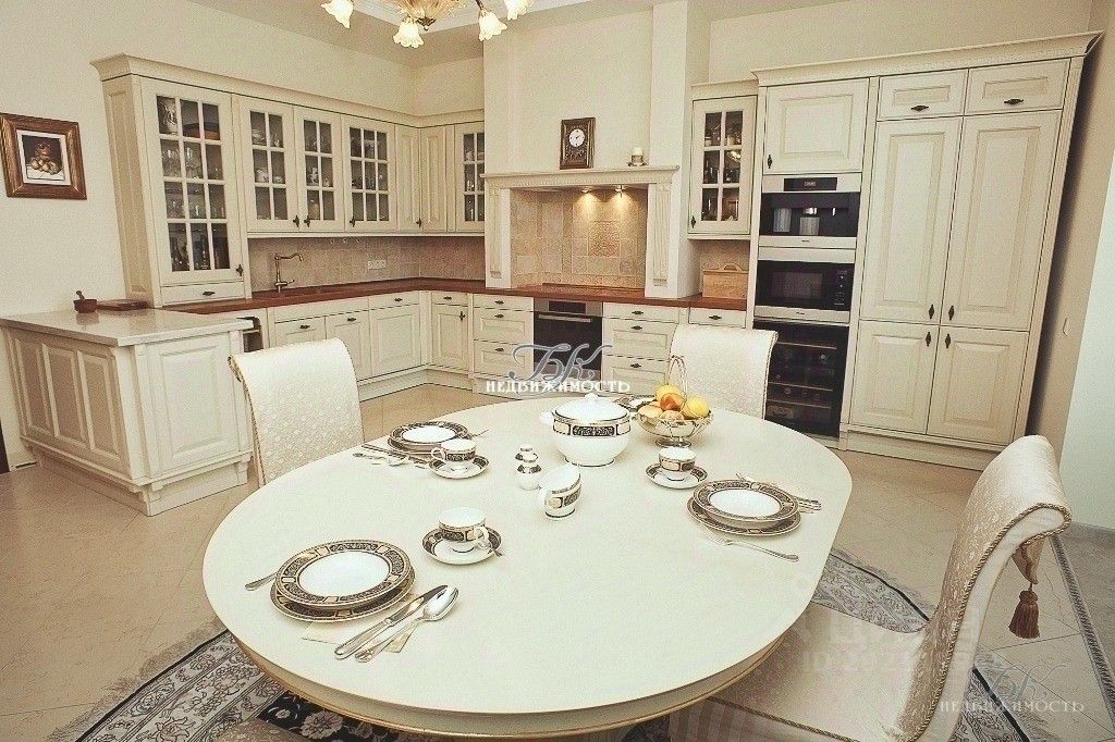 Кухня в классическом стиле с белыми шкафами и деревянной столешницей. Угловая планировка обеспечивает функциональность, а стеклянные дверцы шкафов добавляют элегантности. Центральный круглый стол с изысканной сервировкой и стульями с мягкой обивкой создает уютную атмосферу. На стенах видны текстурированные плитки, а освещение подчеркивает детали интерьера. Использованы светлые тона, что придает пространству ощущение простора и света. Мебель и отделка выполнены из качественных материалов, создавая гармоничное сочетание стиля и комфорта.