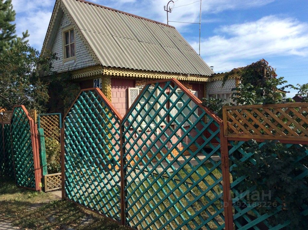 Деревянный дом в дачном стиле с наклонной крышей и мансардным окном. Фасад украшен резными элементами и яркой отделкой, в основном розового и желтого цвета. Забор выполнен из деревянных решеток с комбинированными цветами, создающими уютную атмосферу. Видна небольшая территория перед домом, обрамленная зелеными растениями. Структурные особенности включают простую, но привлекательную архитектуру, характерную для загородных домов. Окна защищены сетками, что добавляет элемент уединения и безопасности.