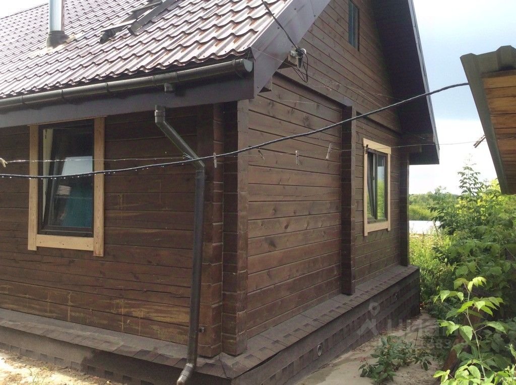 Деревянный дом в современном стиле с крышей из черепицы. Стены выполнены из бревен, отделка натурального дерева придает уютный вид. На фасаде видны окна с деревянными рамами, которые обеспечивают хорошее освещение. Структурные элементы включают выступающие карнизы и наличники вокруг окон. Цветовая гамма тёмная, с оттенками коричневого, что создает гармонию с природой. Внешний вид дома подчеркивает его экологичность и связь с окружающей средой, а также привлекает внимание к качеству используемых материалов.