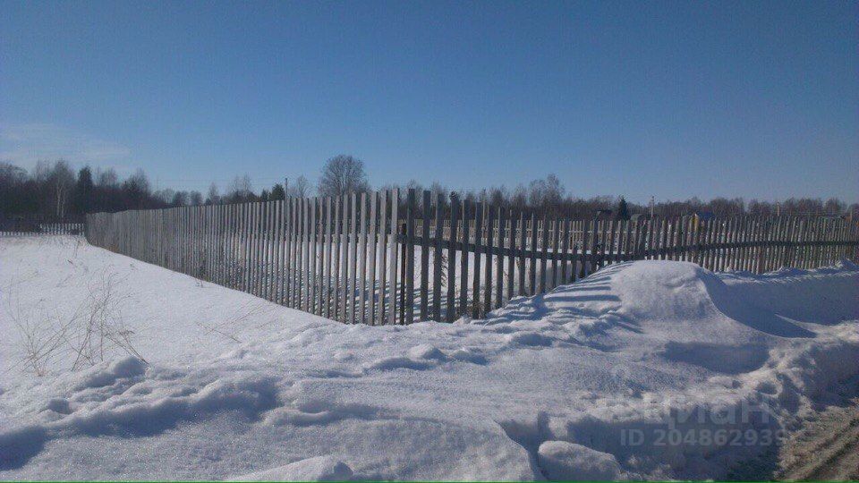 На фотографии видно участок земли, окруженный деревянным забором. Забор выполнен из вертикальных досок, окрашенных в светлый цвет, что придает ему аккуратный вид. На земле лежит снег, создавая контраст с темным деревом забора. Вдали виднеются деревья, а также следы от транспорта на снегу. Участок выглядит просторным и открытым, с ровной поверхностью, что может быть удобно для дальнейшей застройки или использования под другие нужды.