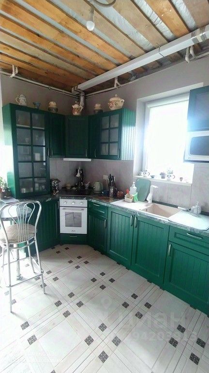 Кухня выполнена в классическом стиле с зелеными деревянными шкафами и стеклянными дверцами. Столешница из светлого камня дополняет общий дизайн. В углу расположена плита и духовка, а рядом находится раковина с современным смесителем. Пол выложен плиткой с геометрическим узором, что придает комнате стильный вид. В помещении много естественного света благодаря окну, которое освещает рабочую зону. Металлический барный стул добавляет современный акцент. На полках видны декоративные элементы, что создает уютную атмосферу.