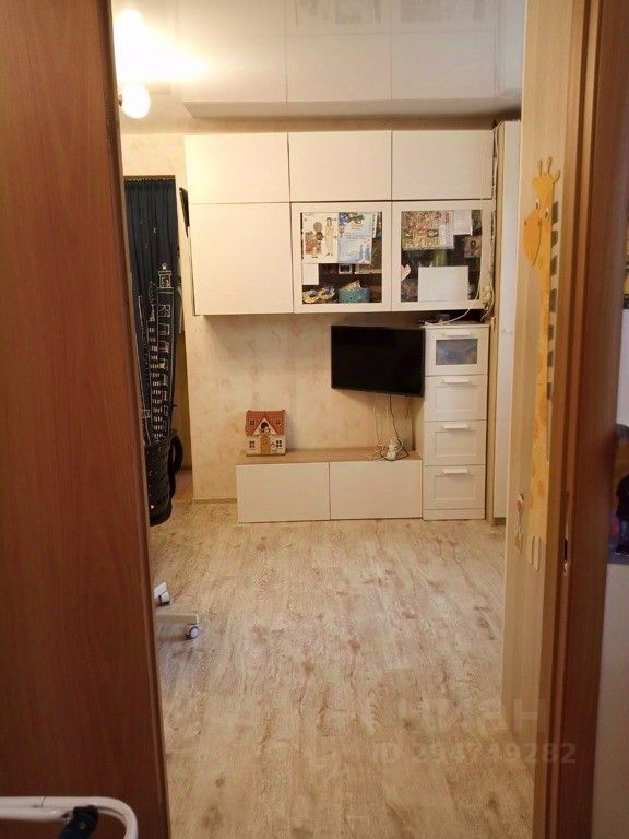 Видна современная комната с минималистичным дизайном. На стенах расположены белые шкафы, в которых видно несколько предметов. Перед ними находится телевизор на стене и низкий комод с ящиками. Пол выполнен из светлого дерева, создающего ощущение простора. В комнате также есть небольшая детская игрушка в виде домика. Освещение мягкое, потолок светлый. Общее оформление интерьера способствует уютной атмосфере, а нейтральные цвета подчеркивают современный стиль.