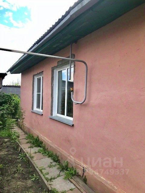 Стена дома выполнена в светло-розовом цвете с гладкой штукатуркой. Окна имеют прямоугольную форму с белыми рамами, придающими свежий и современный вид. Над окнами расположены простые наличники, которые подчеркивают архитектурный стиль. Крыша выполнена из металлического материала, что добавляет прочности конструкции. Вдоль фундамента видны плитки, образующие аккуратную дорожку, окруженную зелеными растениями. Общая эстетика создает уютное и приветливое впечатление, характерное для загородного жилья.