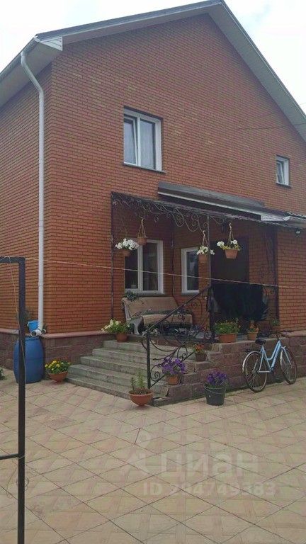 Дом выполнен в современном стиле с кирпичной отделкой, что придаёт ему прочность и уют. На переднем плане видна крытая терраса с металлической перилами и цветочными горшками, создающими атмосферу уюта. Ступени ведут к входной двери, обрамленной окнами, что обеспечивает естественное освещение. Внешние стены окрашены в теплые оттенки, а крыша имеет простой, но элегантный наклон. Полы на террасе выложены плиткой, что добавляет практичности. Рядом с домом расположены элементы декора, такие как велосипед и дополнительные цветочные горшки, подчеркивающие ухоженность пространства.