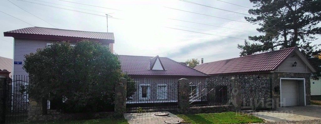 Архитектурный стиль дома сочетает элементы традиционного и современного дизайна, с акцентом на симметричную форму и красную металлическую крышу. Стены выполнены из белого сайдинга, что придает свежий вид. Видна просторная веранда с металлической решеткой, обеспечивающей защиту и открывающей доступ к входу. Основные материалы включают камень и металл, создавая контраст с мягкими зелеными растениями вокруг. Цветовая палитра включает белый, красный и серый, что подчеркивает гармонию с природой и создает уютную атмосферу.