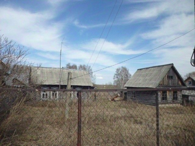 На фотографии представлены деревянные дома в традиционном русском стиле. Архитектурные элементы включают крыши с наклоном и металлические кровли. Структура зданий состоит из бревенчатых стен с большими окнами, что создает уютную атмосферу. Видно, что дома расположены на открытой местности с ограждением. Основные цвета - естественные оттенки дерева и серый металл. Финишные материалы в основном натуральные, что подчеркивает традиционный характер постройки. Пространство вокруг домов выглядит неухоженным, что добавляет исторического контекста к общей картине.