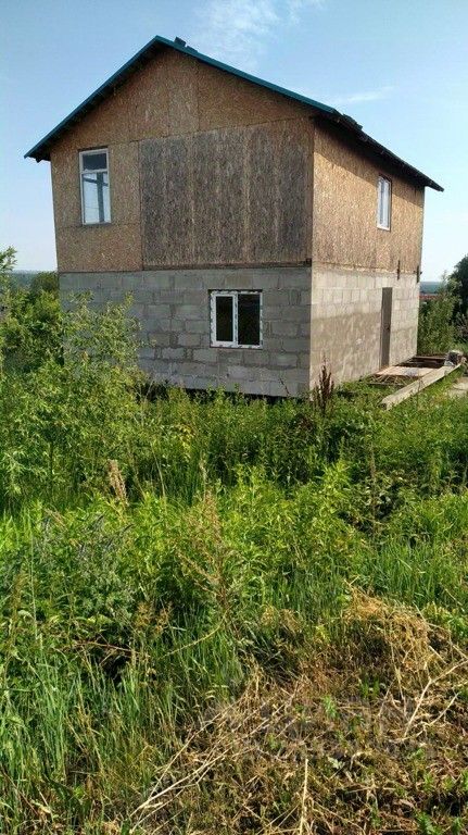Двухэтажный дом с комбинированным фасадом, выполненным из бетонных блоков и деревянных панелей. На верхнем этаже расположены два окна, обеспечивающих естественное освещение. Стены нижнего этажа имеют гладкую текстуру, в то время как верхний этаж отделан фанерой. Крыша с небольшим наклоном, выполненная из металлочерепицы, окрашена в яркий голубой цвет. Вокруг здания растет высокая зелень, создающая естественный ландшафт. Общая структура выглядит простой и функциональной, с акцентом на практичность и доступность материалов.