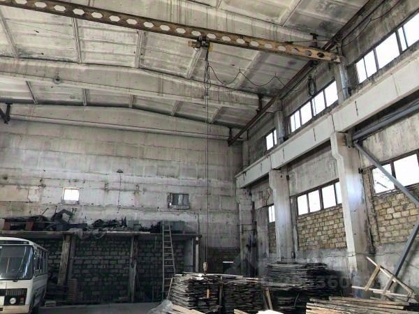 Просторное помещение с высокими потолками и открытой планировкой. Стены выполнены из бетона, с видимыми следами эксплуатации. Внутреннее освещение обеспечивается большими окнами, которые пропускают естественный свет. В центре помещения находится подъемный кран, что указывает на промышленное назначение пространства. На полу расположены штабели материалов, вероятно, строительных плит, а также лестница, ведущая к верхним уровням. Общая атмосфера пространства функциональна и подходит для складирования или производственной деятельности.