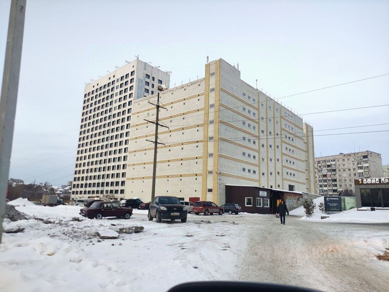 Здание выполнено в современном архитектурном стиле с элементами минимализма. Структура включает многоэтажный корпус с симметричными окнами и простыми линиями. На переднем плане видно парковку с несколькими автомобилями и пешеходной дорожкой. Цветовая палитра фасада включает светлые оттенки, а также контрастные темные окна. Использованы качественные строительные материалы, что придаёт зданию современный вид. Общая планировка предполагает наличие коммерческих помещений на первом этаже, а жилых на верхних уровнях, что создаёт функциональное использование пространства.