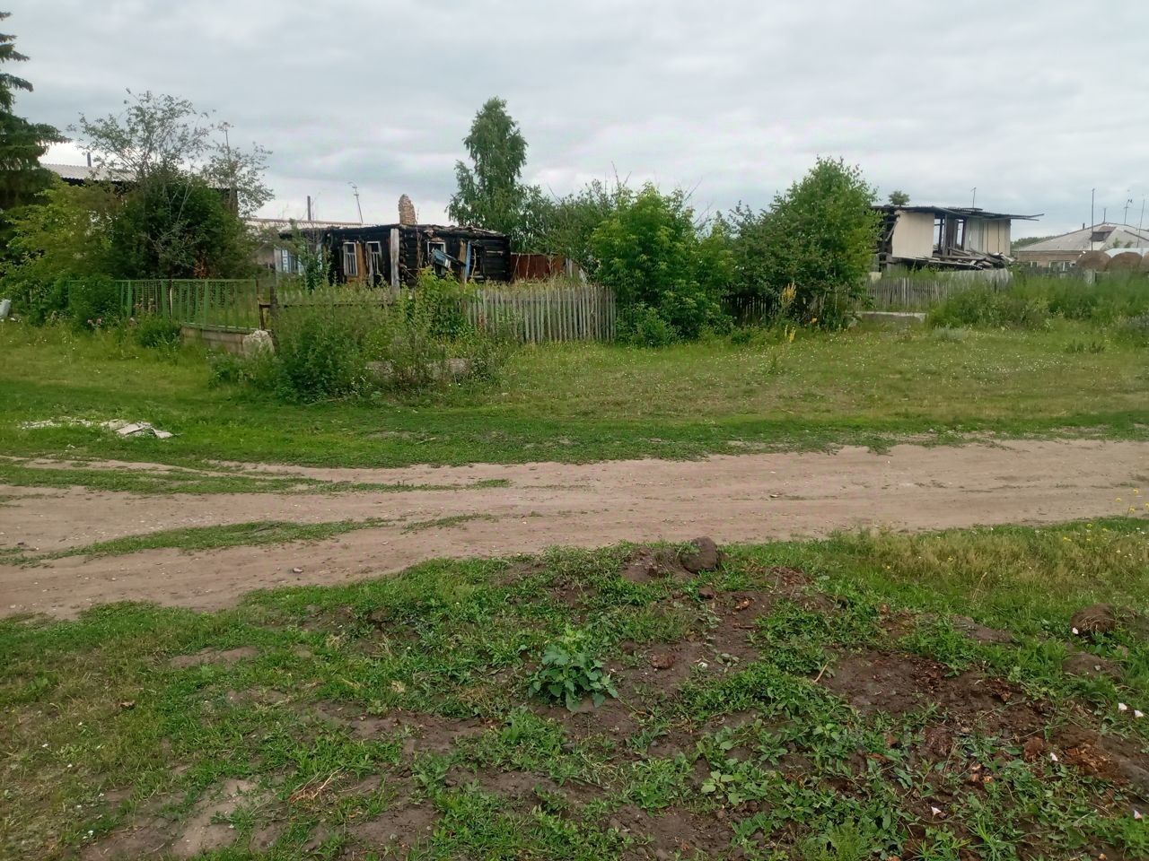 На фотографии представлено открытое пространство с участком земли, окружённым зелеными насаждениями и кустарниками. Видны элементы забора, выполненного из дерева, а также несколько строений, которые имеют простую архитектурную форму. Структура зданий кажется неухоженной, с видимыми следами износа. Основные цвета – это зелёный от травы и кустов, а также коричневые и серые оттенки от строений. Земля на участке выглядит неравномерной, с участками грязи и травы, что создаёт естественный и деревенский ландшафт.
