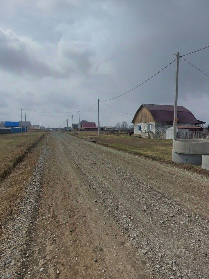 На фотографии представлена сельская местность с дорогой, выложенной гравием. По обеим сторонам дороги расположены дома с различными архитектурными стилями. Некоторые из них имеют деревянные элементы, а другие построены из камня. Крыши домов накрыты шифером или металлом, что придаёт им современный вид. Вдоль дороги стоят столбы с электрическими проводами, создавая ощущение обжитости. Открытое пространство вокруг домов и дороги подчеркивает сельский характер этого района. Цвета в основном нейтральные, с преобладанием серых и коричневых оттенков, что соответствует природному окружению.