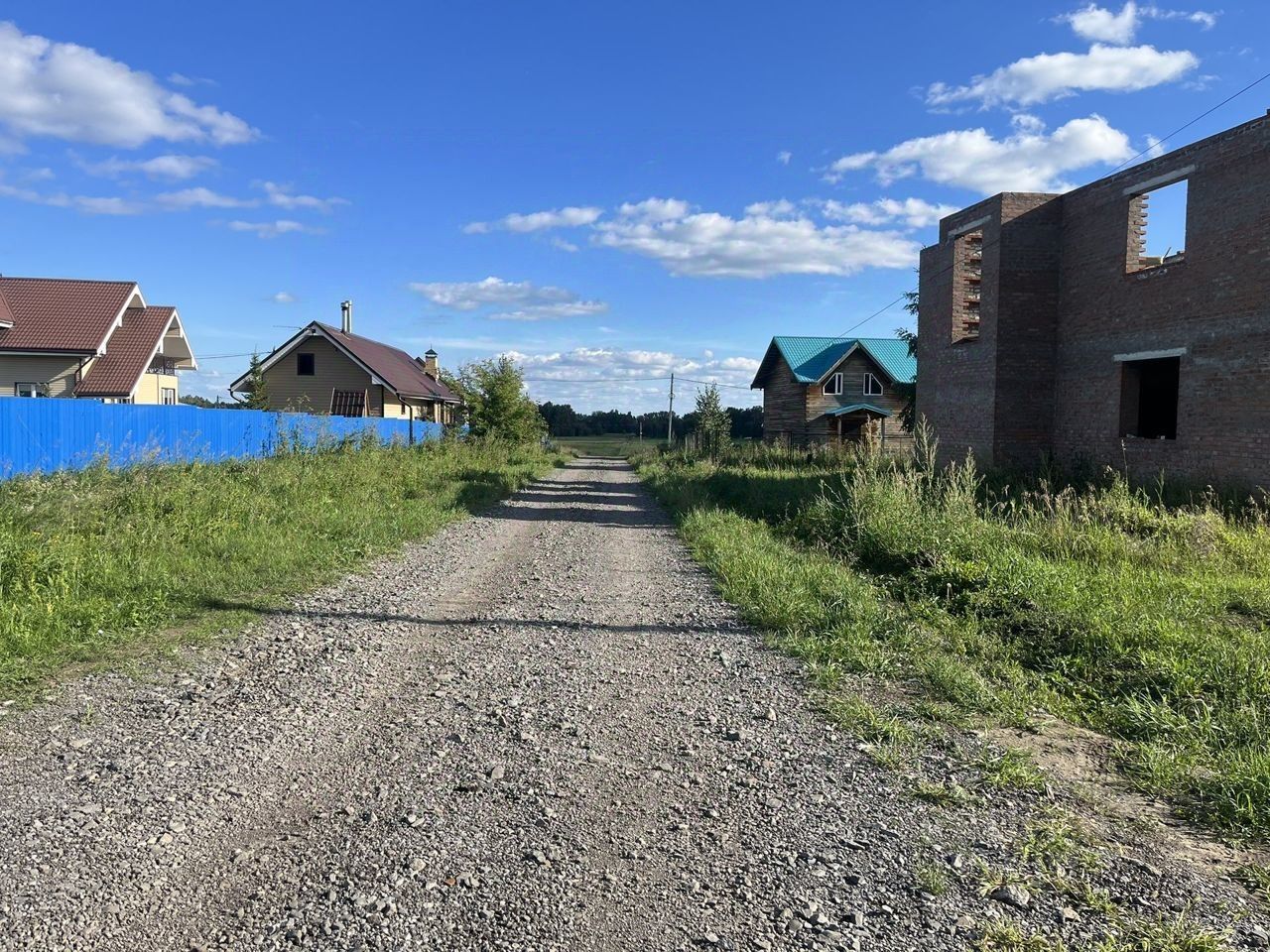 На фотографии видно gravel дорогу, окруженную зелеными травами и кустарниками. С одной стороны дороги расположены дома с разными архитектурными стилями: один из них с крышей из черепицы, другой — с ярко-синей крышей. Строение из кирпича, вероятно, находится на стадии завершения, с открытыми окнами и незавершенной отделкой. Забор из синего металла отделяет участок от дороги, создавая контраст с природным окружением. Пространство выглядит просторным и открытым, что придает ему ощущение уединения и спокойствия.
