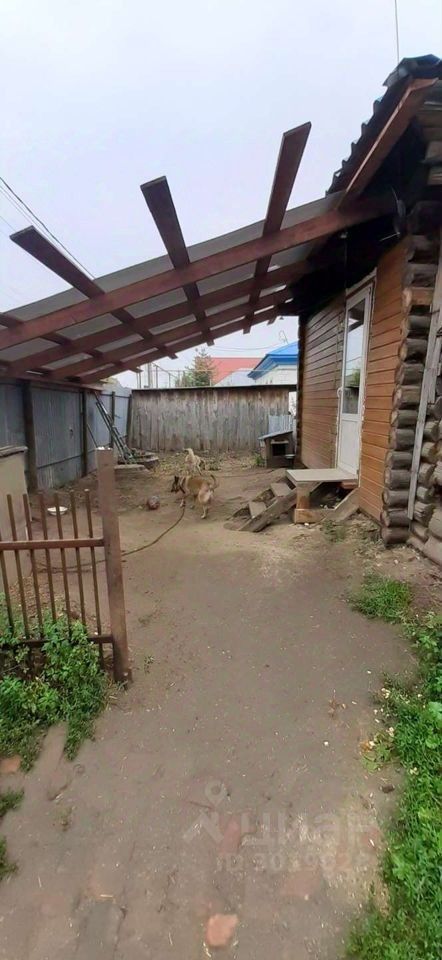Видна деревянная постройка с элементами народного стиля, характерная для загородного жилья. Стены выполнены из бруса, что придаёт уютный вид. На крыше установлен навес из деревянных балок, обеспечивающий защиту от дождя. Перед входом расположены ступени, ведущие к двери. Во дворе виден ухоженный участок с небольшими растениями и травой. На земле лежат небольшие камни, а также видны собаки, что добавляет жизни в пространство. Общая атмосфера спокойная и домашняя, акцент на природные материалы и простоту дизайна.