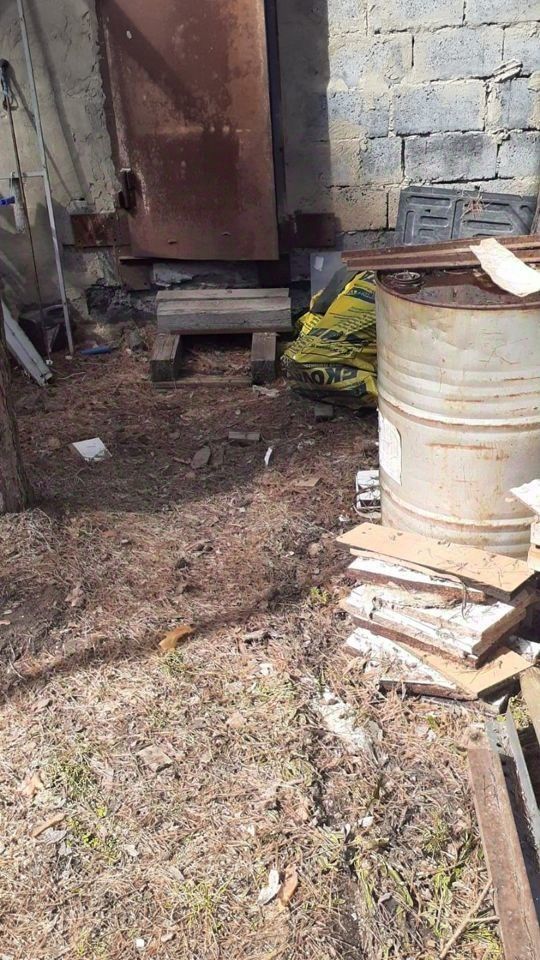 Видно пространство с грубой отделкой, где преобладают бетонные и металлические элементы. На заднем плане расположена старая металлическая дверь, имеющая следы коррозии. Пол выложен из деревянных поддонов и обрезков, создавая неравномерную поверхность. В углу стоит металлическая бочка, рядом с ней находятся мешки с материалами. Окружающая среда выглядит запущенной, с остатками строительных материалов и природным мусором. Цветовая палитра включает серые, коричневые и металлические оттенки, что придаёт пространству индустриальный вид.