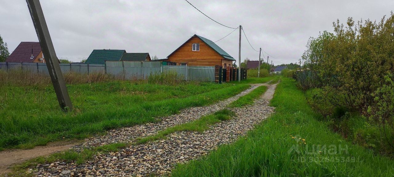 На фотографии виден загородный участок с гравийной дорогой, окруженный зелеными травами и кустарниками. Вдалеке расположены деревянные дома с крышей из металлочерепицы, выполненные в простом архитектурном стиле. Один из домов имеет светлую деревянную отделку, а другой - темно-зеленую крышу. Заборы из металлических панелей обрамляют участок, создавая уединение. Дорога имеет неровную поверхность, что характерно для загородных местностей. Общее впечатление создает атмосферу спокойствия и близости к природе.