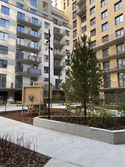 Видна современная архитектура с элементами минимализма, включая прямые линии и большие окна. Структурные особенности включают балконы с металлическими перилами и кирпичные стены. Изображена общая зона с озеленением, где расположены скамейки и детская площадка. Основные характеристики включают ухоженные клумбы, асфальтированные дорожки и аккуратно подстриженные деревья. Материалы включают бетонные и кирпичные конструкции, а цветовая гамма состоит из светлых и нейтральных тонов, создающих гармоничное восприятие пространства.
