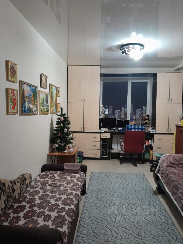 Комната оформлена в современном стиле с элементами минимализма. На стенах висят картины в рамках, добавляющие уют. Видны встроенные шкафы из светлого дерева, что создает ощущение простора. Основное внимание привлекает рабочая зона с компьютером и стулом, расположенная у окна. В комнате присутствует небольшой новогодний елка, что создает праздничную атмосферу. Пол покрыт мягким ковром с узором, а мебель оформлена в нейтральных тонах. Освещение обеспечивается стильной потолочной люстрой, добавляющей теплый свет в пространство.