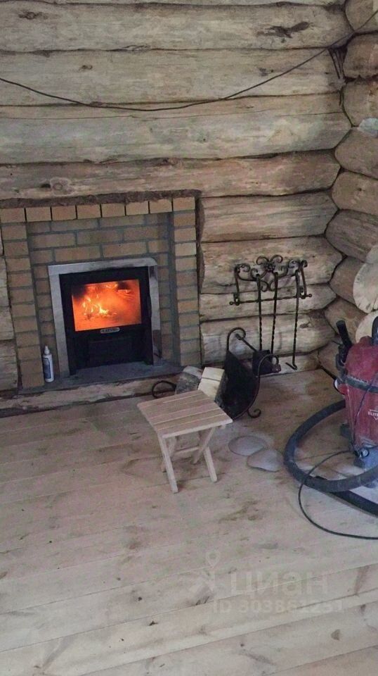 В интерьере представлено помещение в стиле загородного дома с элементами деревянной архитектуры. Стены выполнены из бревен, что придает пространству уютную атмосферу. В центре находится современный камин с кирпичной отделкой, создающий теплый и комфортный уголок. Полы покрыты светлым деревом, что визуально расширяет пространство. Видны также небольшая деревянная скамейка и несколько предметов, связанных с обустройством, что подчеркивает функциональность и домашний уют. Цветовая палитра теплых тонов гармонично сочетается с природными материалами, создавая атмосферу спокойствия.