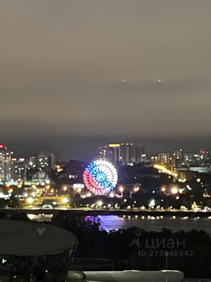 На фотографии виден городской пейзаж с ярко освещённым колесом обозрения, которое выделяется своими красочными огнями. Архитектурный стиль зданий на заднем плане варьируется от современных высоток до более традиционных конструкций. Структурные элементы включают в себя мост и набережную, которые создают гармоничное сочетание с городской средой. Основные характеристики этого района подчеркиваются ночным освещением, создающим атмосферу уюта и динамики. Цветовая палитра включает в себя тёплые и холодные оттенки, что придаёт изображению живость и привлекательность.