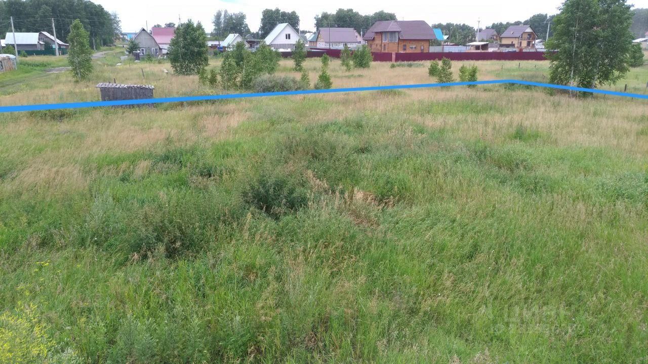 На фотографии видно обширное открытое пространство с высокой травой и редкими кустарниками. Земельный участок имеет неровный рельеф, с небольшими возвышенностями и впадинами. Вдали видны строения, включая дома с крышей из металлочерепицы и заборы. Цвета преобладают зеленые оттенки травы и коричневые тона почвы. Участок окружен небольшими деревьями, создающими ощущение уединенности. Общая атмосфера пространства спокойная и естественная, что делает его привлекательным для дальнейшего использования или строительства.