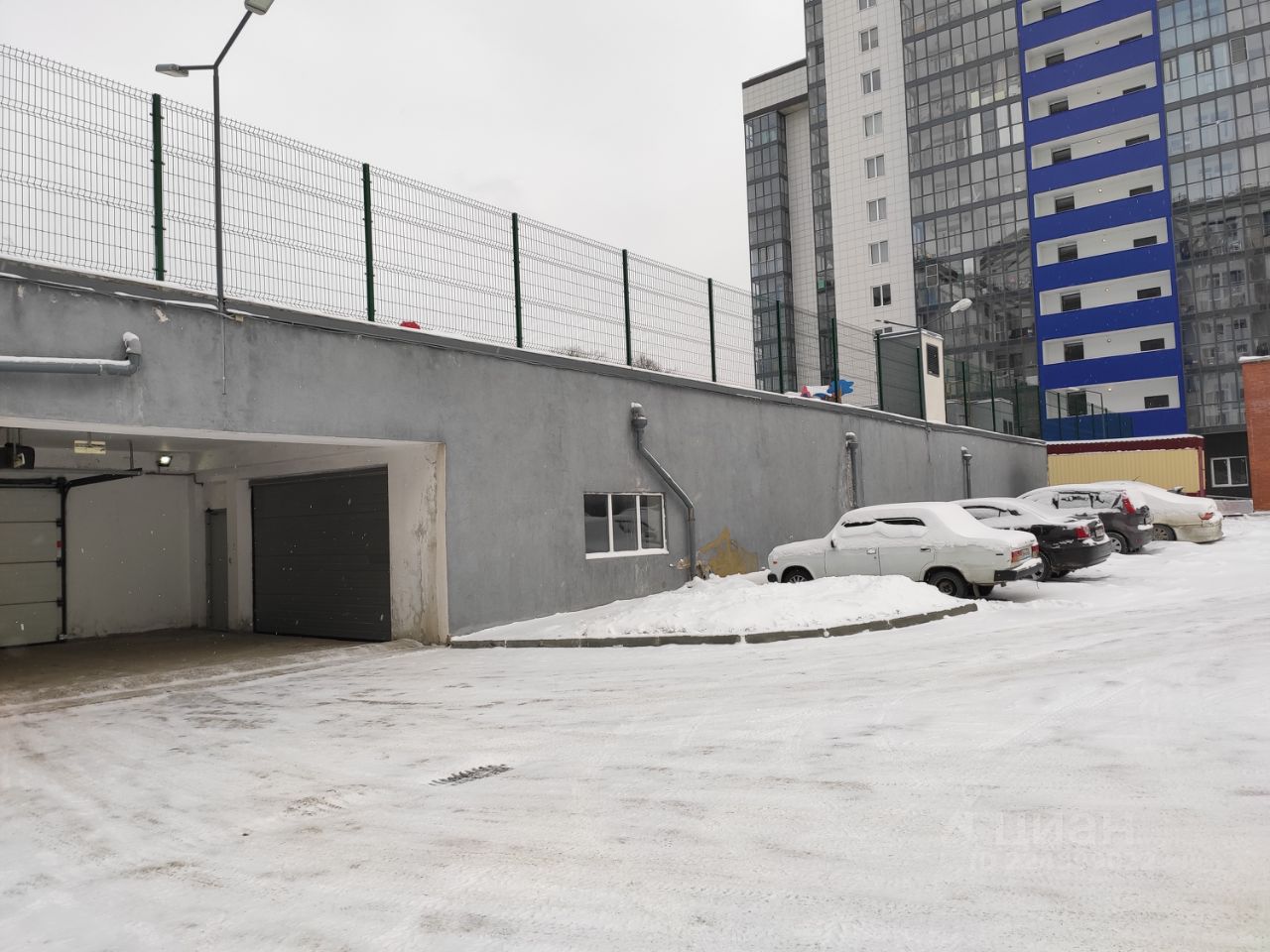Видна парковка с асфальтированным покрытием, окруженная серыми стенами. Вход в подземный гараж оформлен автоматическими воротами. Стены выполнены из бетона, окрашенного в серый цвет. На парковке припаркованы несколько автомобилей, часть из которых покрыта снегом. Слева расположены окна, обеспечивающие естественное освещение. Высокая металлическая ограда с зелеными прутьями возвышается над стеной, создавая ощущение безопасности. Освещение представлено несколькими фонарями, установленными на столбах. Общая атмосфера пространства выглядит функционально и современно.