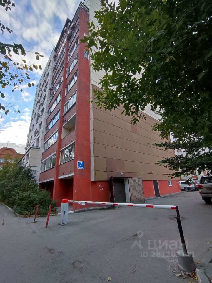 Здание выполнено в современном архитектурном стиле с элементами минимализма. Фасад комбинирует красные и бежевые панели, создавая контраст. Видна многоэтажная структура с большими окнами, обеспечивающими естественное освещение. На первом этаже расположены входные группы и гаражные ворота. Основные материалы – бетон и стекло, отделка аккуратная и современная. Цветовая гамма включает теплые и нейтральные оттенки, что придает зданию дружелюбный и привлекательный вид. Входная зона ограничена шлагбаумом, что подчеркивает безопасность и приватность.