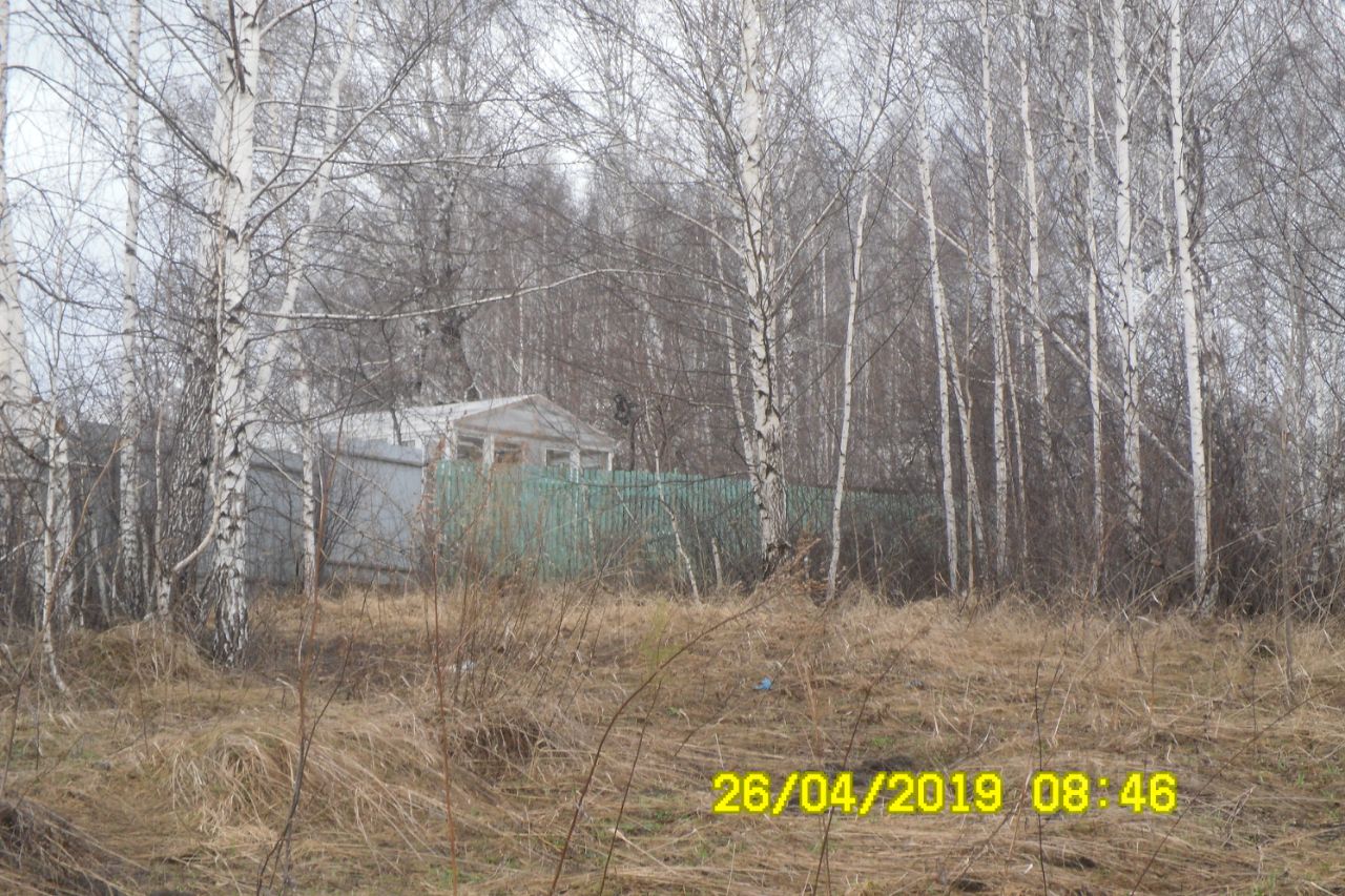 На фотографии виден небольшой дом с простым архитектурным стилем, выполненный из светлого материала. Структурные особенности включают плоскую крышу и окна, расположенные по бокам. Вокруг дома находится забор из металлической сетки, который частично скрывает участок. Основные характеристики пространства включают неухоженный участок с травой и редкими кустами. Цветовая палитра доминирует в светлых и серых тонах, а отделка выглядит простой и функциональной, что подчеркивает сельский стиль жизни.