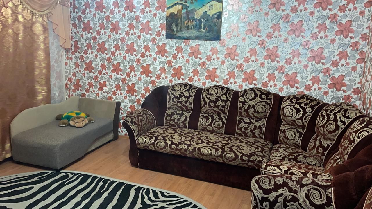 Комната оформлена в уютном стиле с яркими акцентами. На стенах расположены обои с крупными цветами, создающими атмосферу тепла и уюта. Основной элемент мебели — большой диван с богатой текстурой и узором, выполненный в темных тонах. Рядом стоит небольшой диван с гладкой обивкой, на котором лежит игрушка. На полу лежит ковер с зебровым принтом, добавляющий динамичности в интерьер. Окна украшены легкими шторами, что позволяет мягкому свету проникать в пространство, подчеркивая его уют и комфорт.