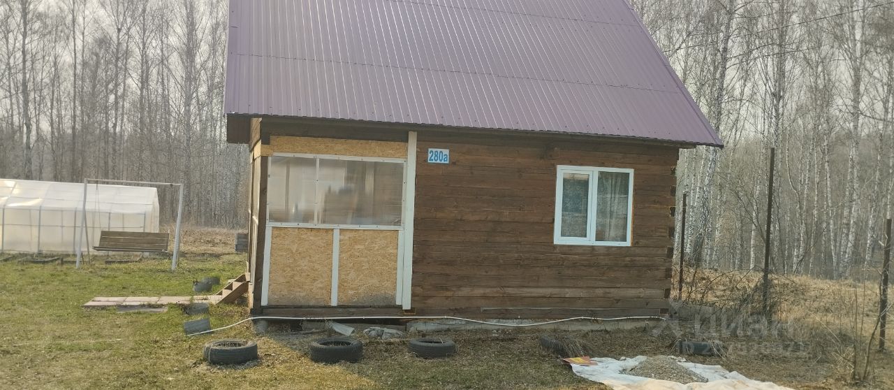Строение выполнено в традиционном стиле с деревянными стенами и крышей из металлочерепицы. На фасаде расположены окна с белыми рамами, а также двери, обрамленные деревянными панелями. Структура имеет простой, но функциональный дизайн, с небольшим крыльцом. Основные материалы — дерево и металл, что придаёт дому естественный вид. Цветовая гамма включает теплые коричневые оттенки дерева и серый металл крыши. Пространство вокруг дома организовано с использованием камней и старых автомобильных шин, создавая уютную атмосферу загородного жилья.
