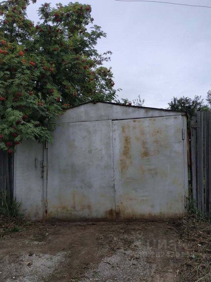 На фотографии изображен металлический гараж с большими распашными воротами. Стены гаража выполнены из металла, имеют ржавые пятна и следы износа, что указывает на длительное использование. Ворота закрыты, но их конструкция предполагает достаточную ширину для въезда автомобиля. Перед гаражом находится небольшая площадка, выровненная с грунтом, с редкими травами. Слева виден куст с яркими ягодами, что добавляет немного зелени к общему виду. Общая атмосфера места создает впечатление утилитарного пространства, предназначенного для хранения транспортных средств или инструментов.