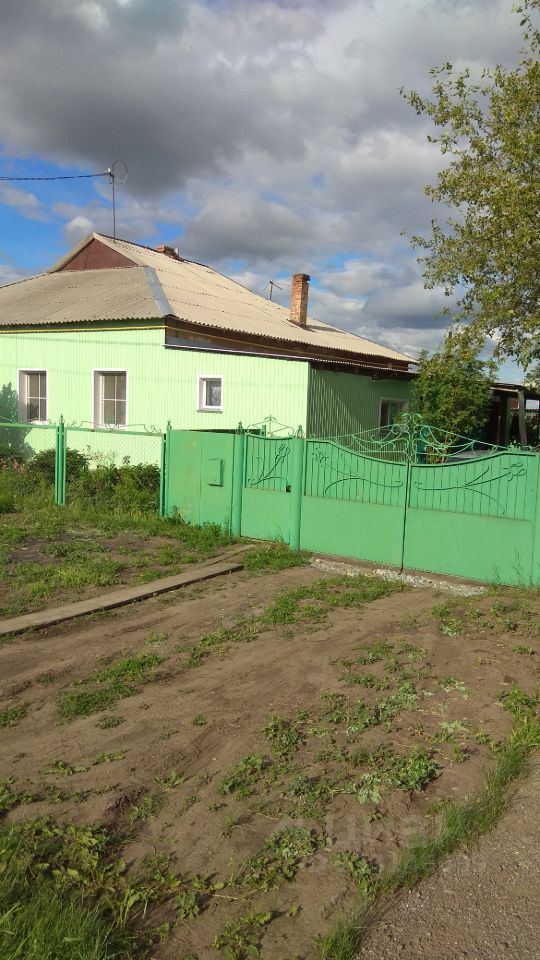 Дом в стиле дачи с яркой зеленой обшивкой и крышей с наклоном. Окна имеют простую прямоугольную форму, что придает зданию традиционный вид. Входные ворота выполнены из металла и окрашены в тот же зеленый цвет, что и фасад, с изящными элементами кованого железа. Перед домом видна ухоженная территория с небольшим склоном и участками земли, поросшими травой. На крыше заметна металлическая черепица, а также антенна, что указывает на наличие современных удобств. Общая атмосфера пространства создает ощущение уюта и спокойствия.