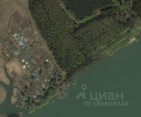 На изображении виден участок земли, окруженный водой и лесом. Архитектурный стиль строений разнообразен, от простых до более сложных форм. Структурные элементы включают несколько зданий, расположенных в ряд, с различными крышами и цветами. Основные характеристики включают открытые пространства и доступ к воде. Видимые материалы варьируются от дерева до металла, с различными отделками. Цвета зданий разнообразные, с преобладанием нейтральных тонов, что создает гармоничный облик с природным окружением.