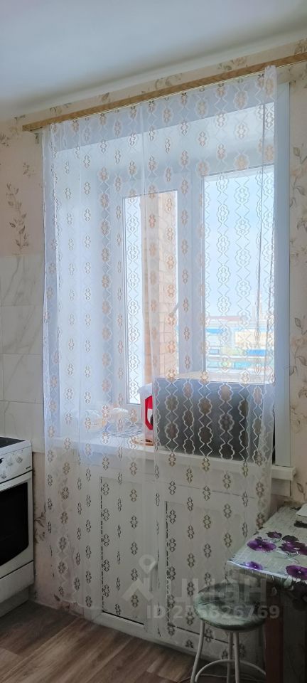 Кухня оформлена в светлых тонах с нежными цветочными узорами на обоях. Окно, обрамленное прозрачной занавеской, позволяет проникать естественному свету, создавая уютную атмосферу. В углу расположена плита, выполненная в классическом стиле, а рядом — белый кухонный шкаф с гладкими фасадами. Пол покрыт светлым ламинатом, а стол с цветочной скатертью добавляет яркие акценты. Пространство организовано функционально, с акцентом на комфорт и стиль, что делает его привлекательным для приготовления пищи и общения.